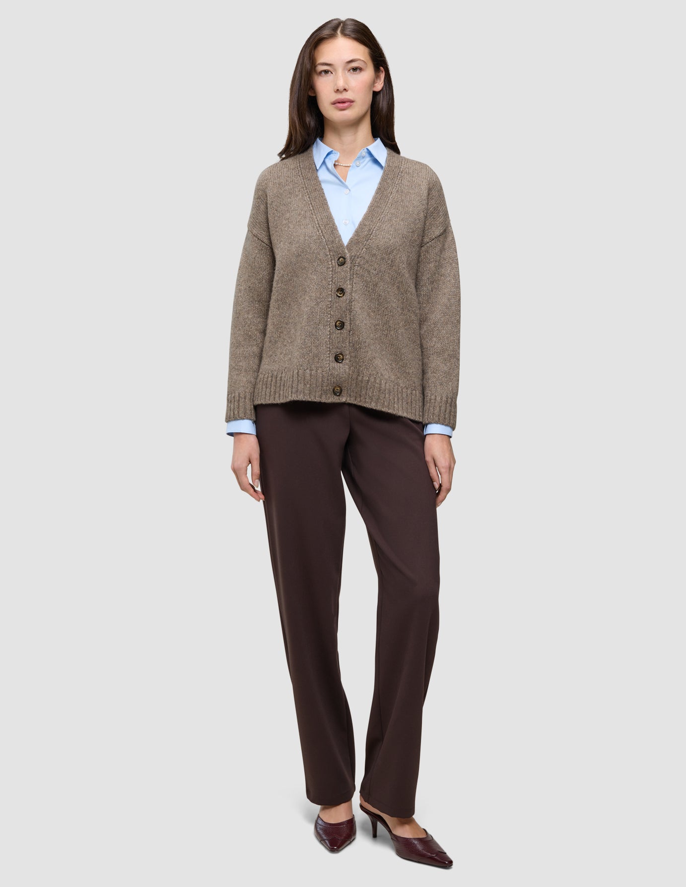 Eine Frau, die eine taupefarbene, melierte, grob gestrickte Damen-Strickjacke aus weicher Wolle über einem hellblauen Hemd mit Kragen trägt, dazu eine dunkelbraune Hose und passende Pumps mit spitzer Zehenpartie.